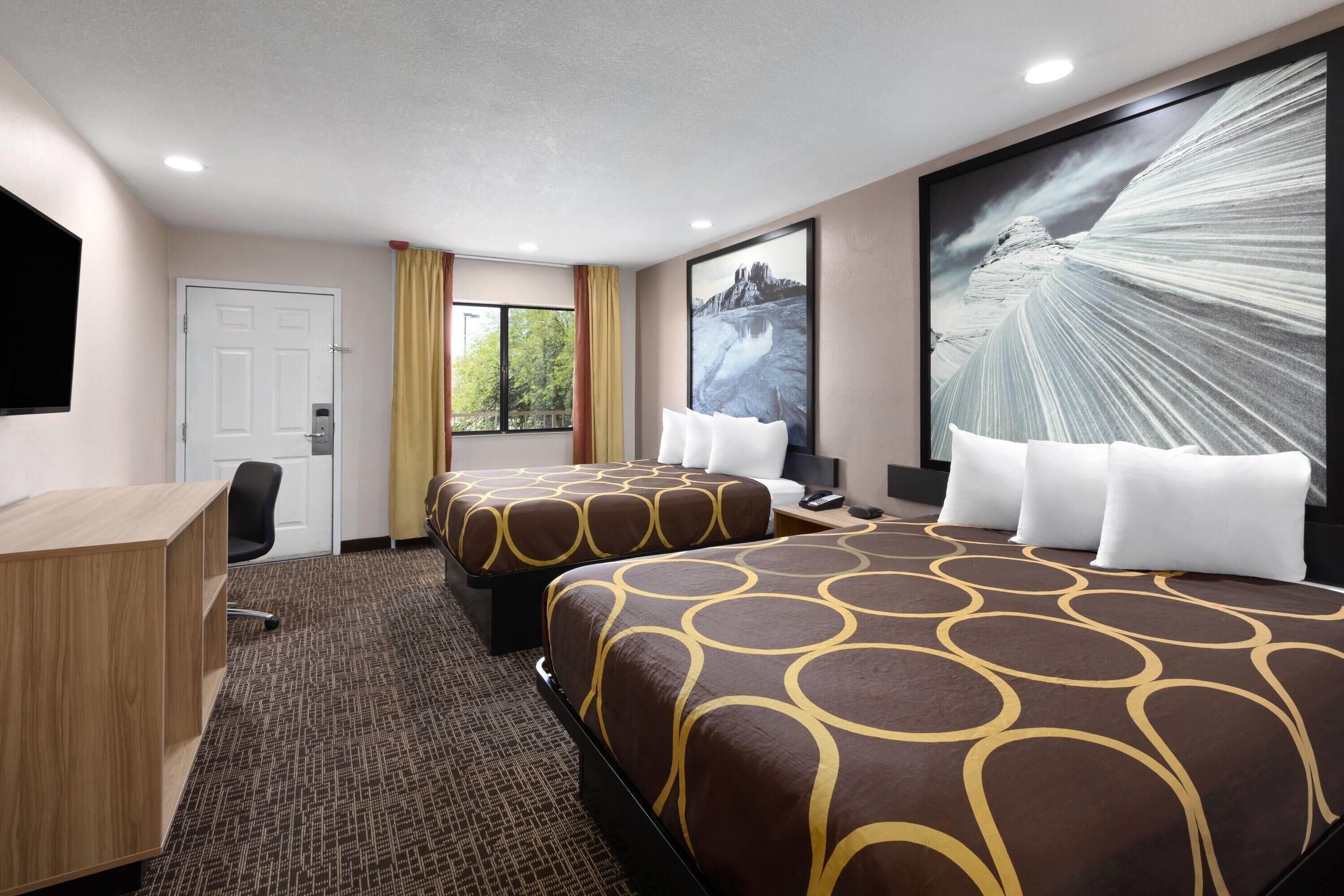 A hotel room with two beds, a desk, a chair, a window with curtains, and a large framed picture on the wall.
