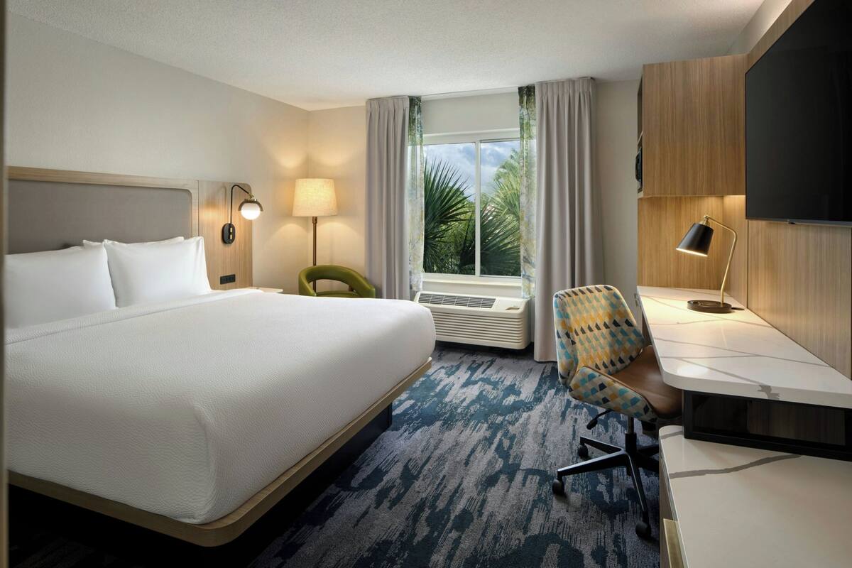 Ein Hotelzimmer mit einem großen Bett, einem Schreibtisch mit Stuhl, einem Fernseher und einem Fenster mit Vorhängen.