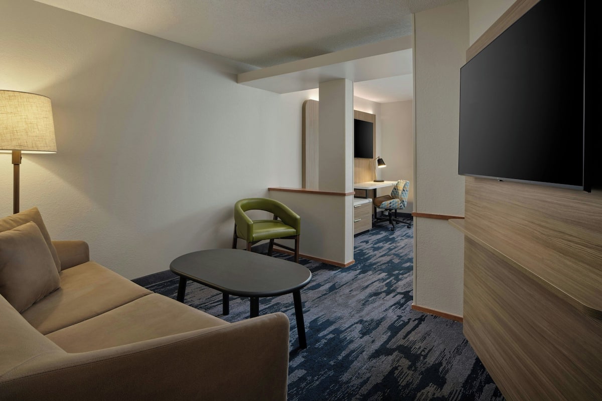 Ein Hotelzimmer mit einer Sitzecke, einem kleinen Tisch, einem grünen Sessel, einem Flachbildfernseher und einem Schreibtisch mit Lampe.