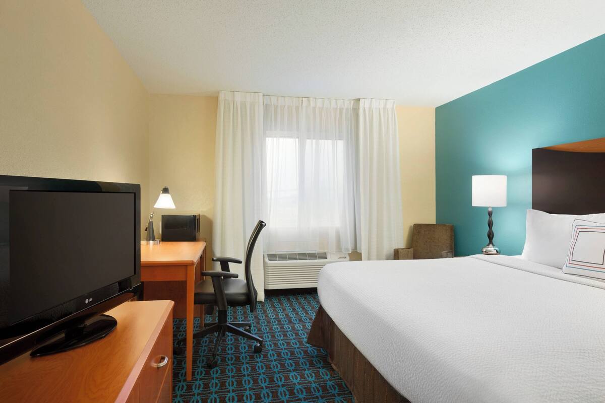 Une chambre d’hôtel avec un lit, un bureau avec une télévision, une chaise, une lampe et une fenêtre avec des rideaux.