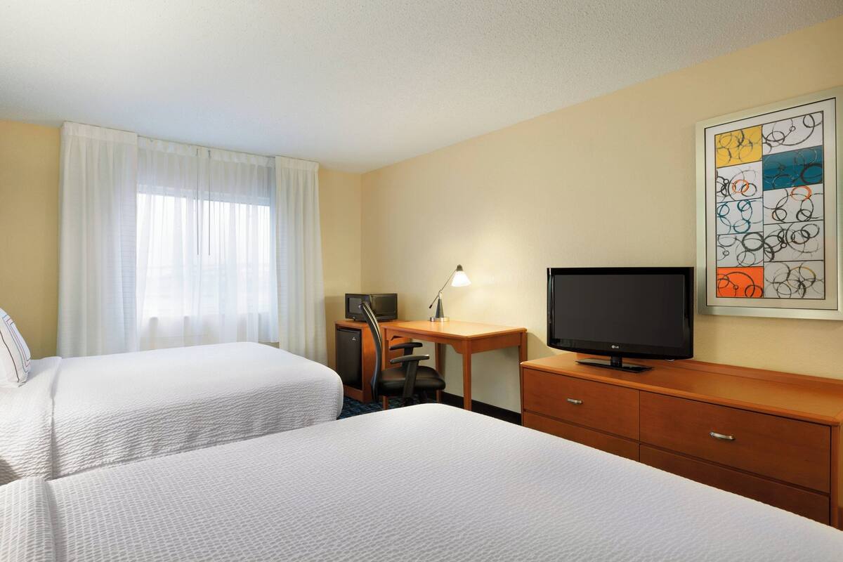 Une chambre d’hôtel avec deux lits, un bureau équipé d’un ordinateur et d’une lampe, une télévision, et une œuvre d’art abstraite encadrée accrochée au mur.