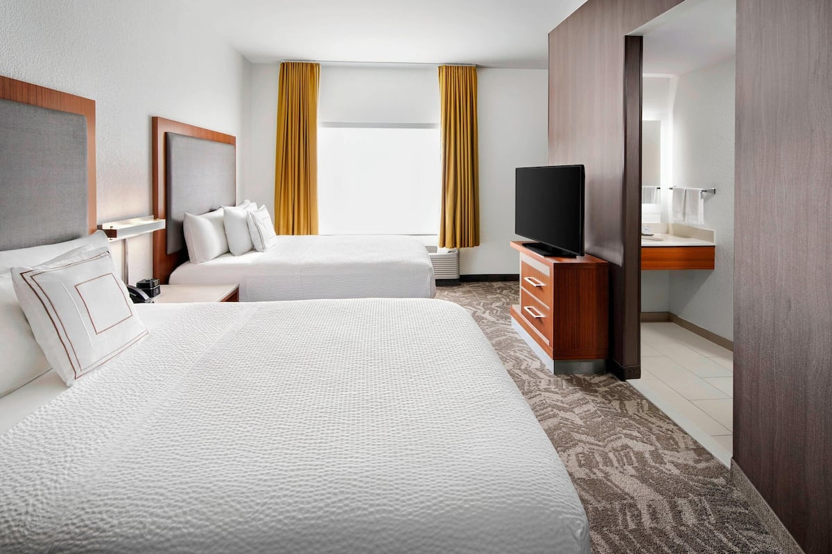 Habitación de hotel con dos camas, un televisor, un escritorio y un baño visible a través de una puerta abierta.