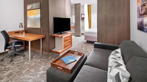 Quarto de hotel com escrivaninha de madeira, televisão de tela plana, sofá e mesa de apoio com um livro.