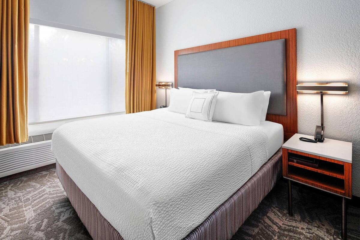 Una habitación de hotel con una cama grande, una mesita de noche con lámpara y una ventana con cortinas.