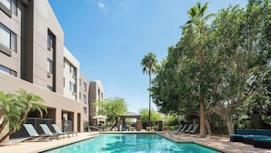 Piscina com espreguiçadeiras e palmeiras, cercada por um edifício com várias janelas e uma área de estar coberta.
