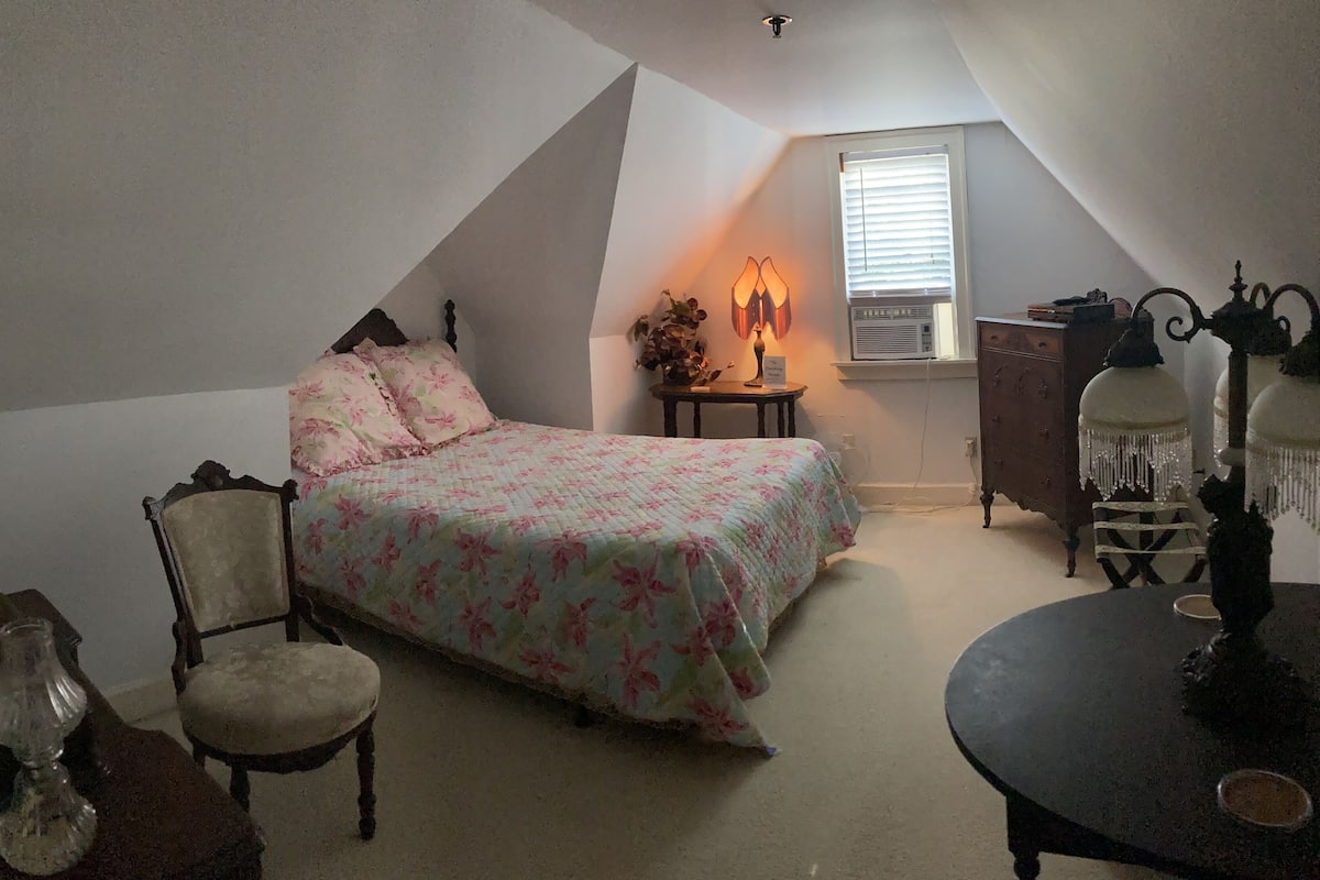 A bedroom with a sloped ceiling, a bed with floral bedding, a chair, a small table with a lamp, and a dresser.