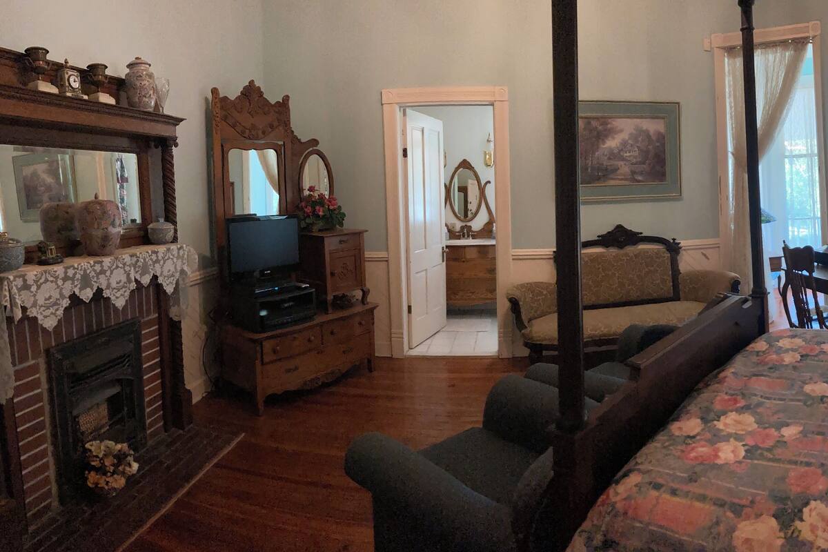A traditional bedroom with a four-poster bed, a fireplace, a television, and a door leading to another room.