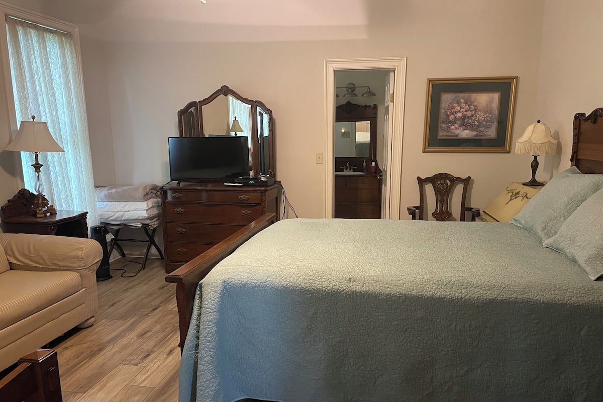 A bedroom with a bed, a dresser, a television, a chair, and a door leading to another room.