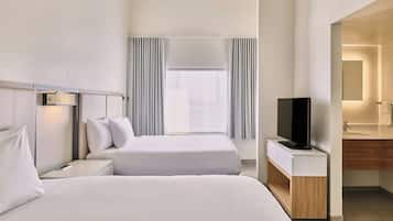 A hotel room with two beds, a TV, a bedside table, a window with curtains, and a bathroom with a mirror and sink.
