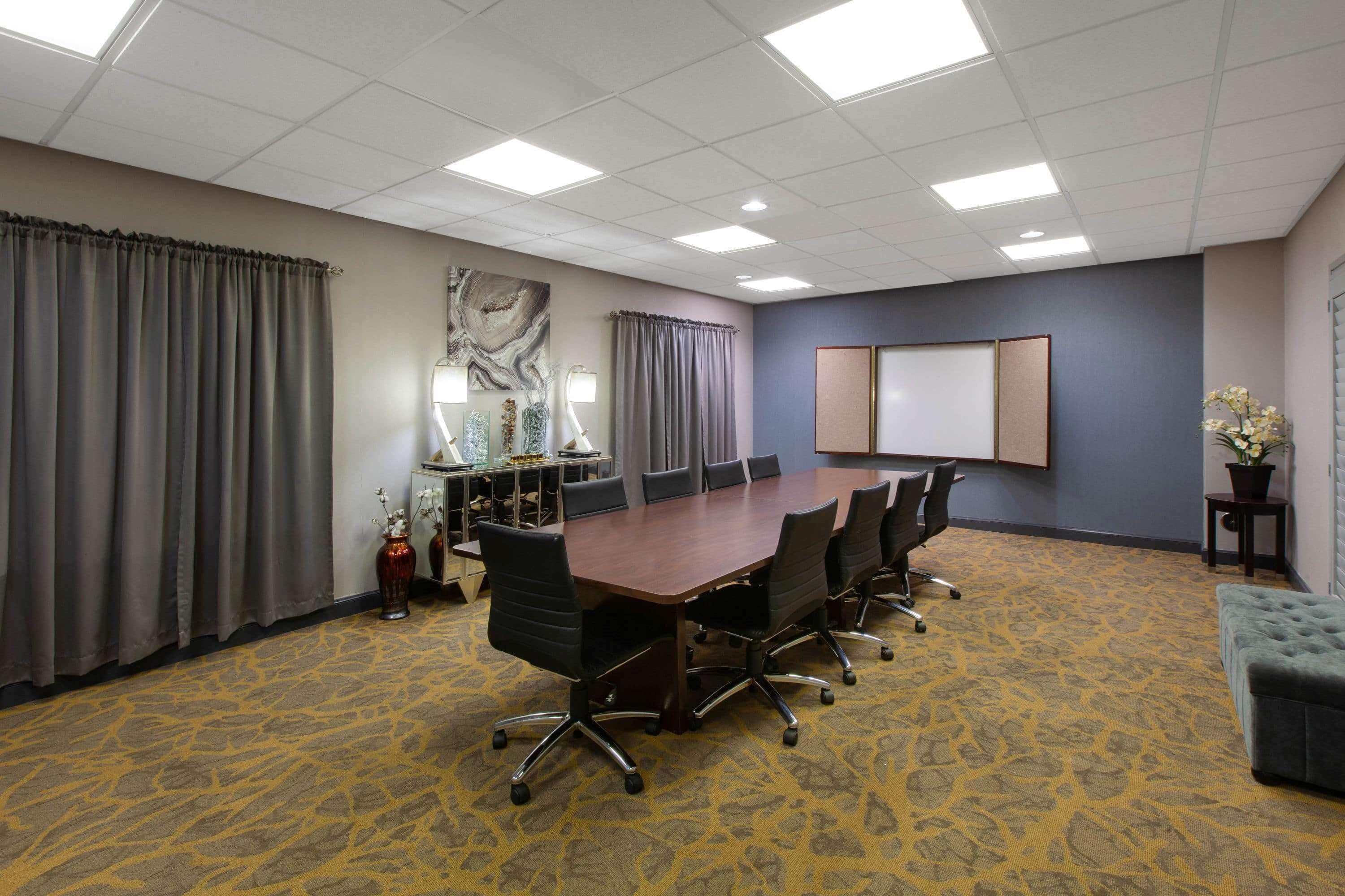 Un salón de conferencias con una gran mesa, sillas, piso alfombrado, cortinas, una planta en maceta y una pizarra.