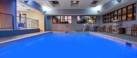 Uma piscina interna com água azul cristalina, uma escadaria que leva a uma varanda e uma área de estar com cadeiras e mesas.