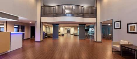 A spacious lobby with a wooden floor, a reception desk, a seating area, and a mezzanine level with a railing.