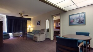 Une chambre d’hôtel équipée d’un canapé, d’un fauteuil, d’une table et de chaises. On y trouve également un ventilateur de plafond, un tableau au mur et une fenêtre avec des rideaux.