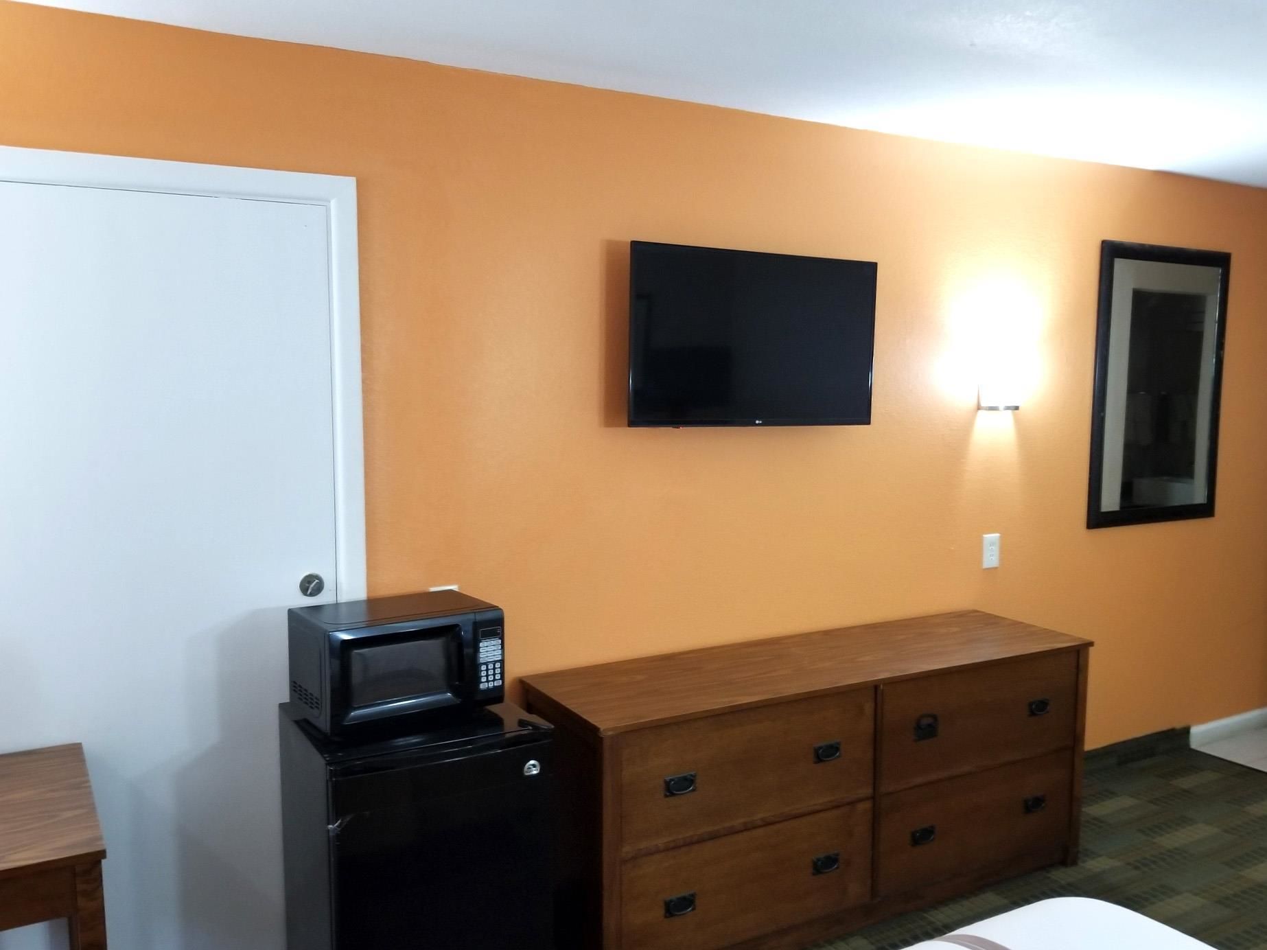 A room with a white door, a black microwave, a wooden dresser with drawers, a flat-screen TV mounted on the wall, and a mirror.