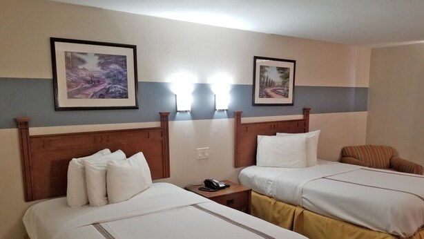 Une chambre d’hôtel avec deux lits, chacun doté de draps blancs et d’une tête de lit en bois. Les murs sont ornés de tableaux encadrés, il y a une table de chevet avec un téléphone et une chaise à rayures.