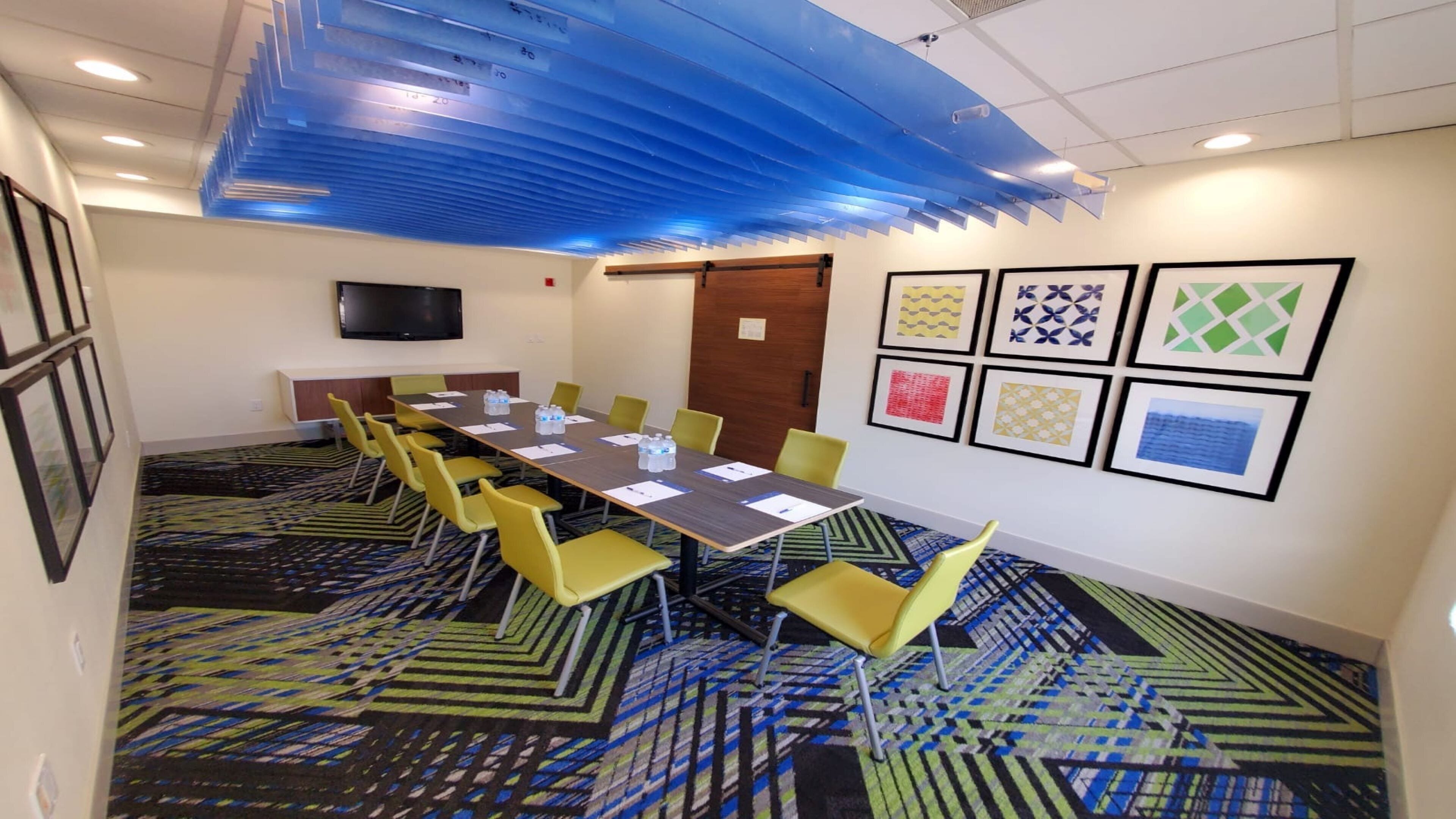 A conference room with a long table, yellow chairs, a flat-screen TV, and patterned carpet.