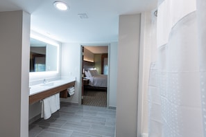A modern bathroom with a large mirror, a wooden vanity, a shower area, and a visible bedroom with a bed and nightstand.