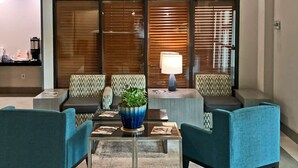 A modern lounge area with teal armchairs, a glass coffee table with a blue vase, and a wooden partition with blinds.