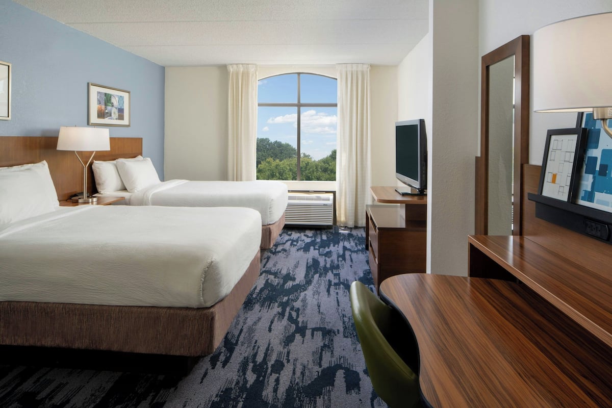A hotel room with two beds, a desk, a TV, and a large window with a view of greenery.