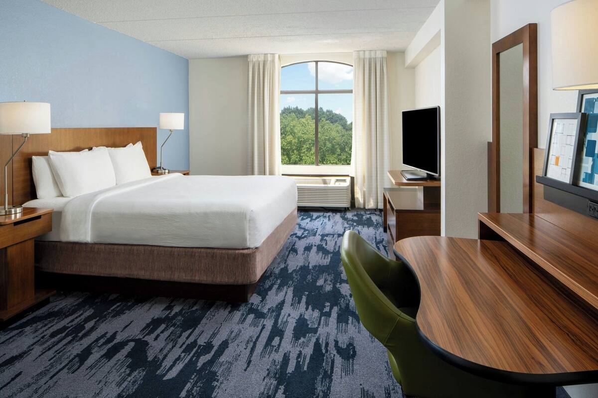 A hotel room with a large bed, a desk, a chair, a TV, and a window with a view of trees.