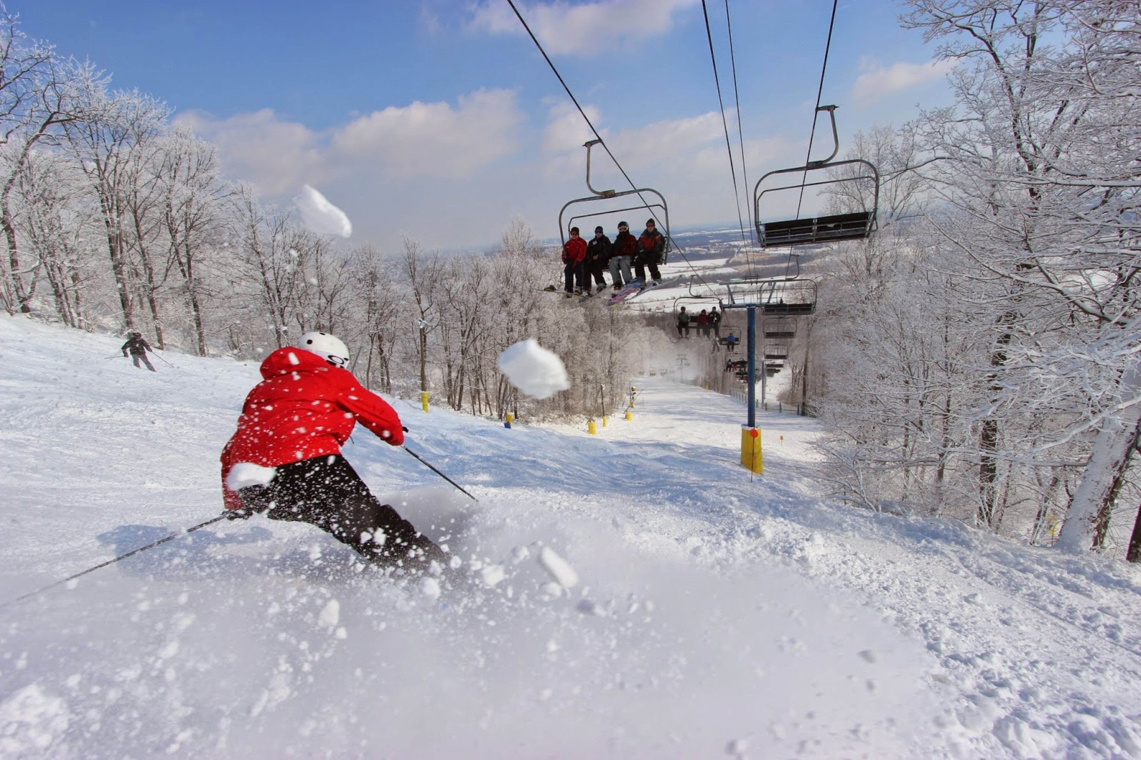 Eine Skipiste mit einer Person beim Abfahren, eine Sesselbahn mit Personen sowie schneebedeckte Bäume.