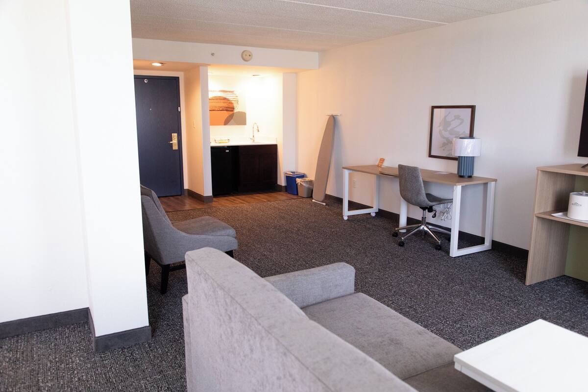 A hotel room with a grey sofa, a small white table, a desk with a chair, a TV, and a bathroom visible through an open door.