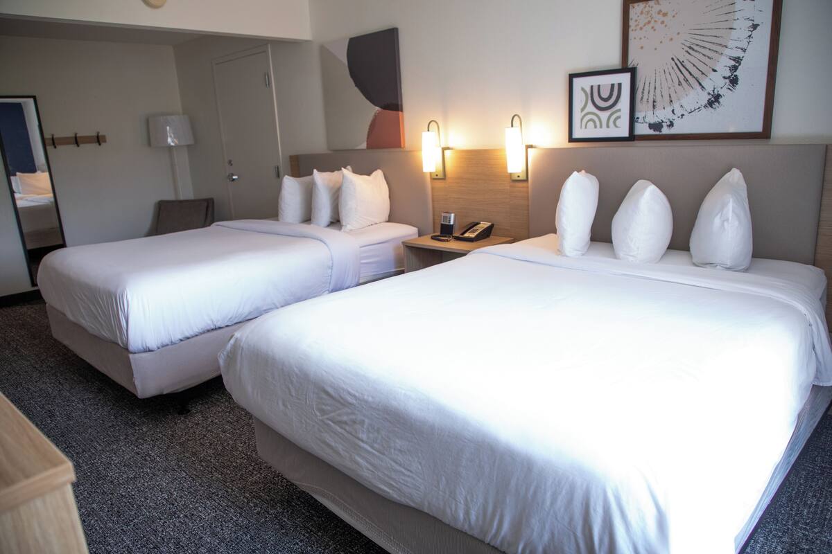A hotel room with two beds, a bedside table with a phone, a lamp, and wall art.
