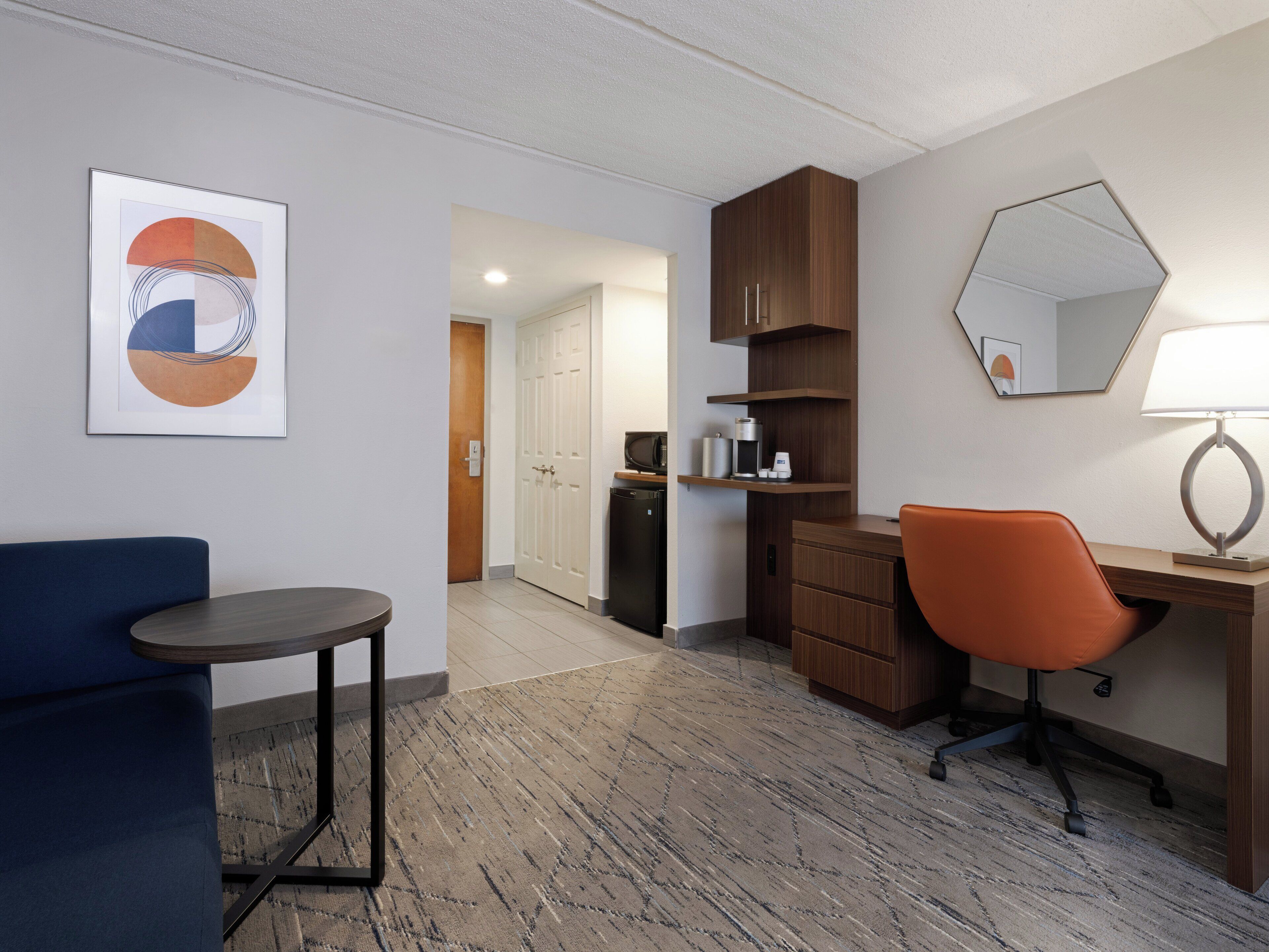 Habitación de hotel con escritorio, silla y una pequeña mesa. Hay un microondas, cafetera y un armario. Un cuadro enmarcado está en la pared, y un espejo encima del escritorio.