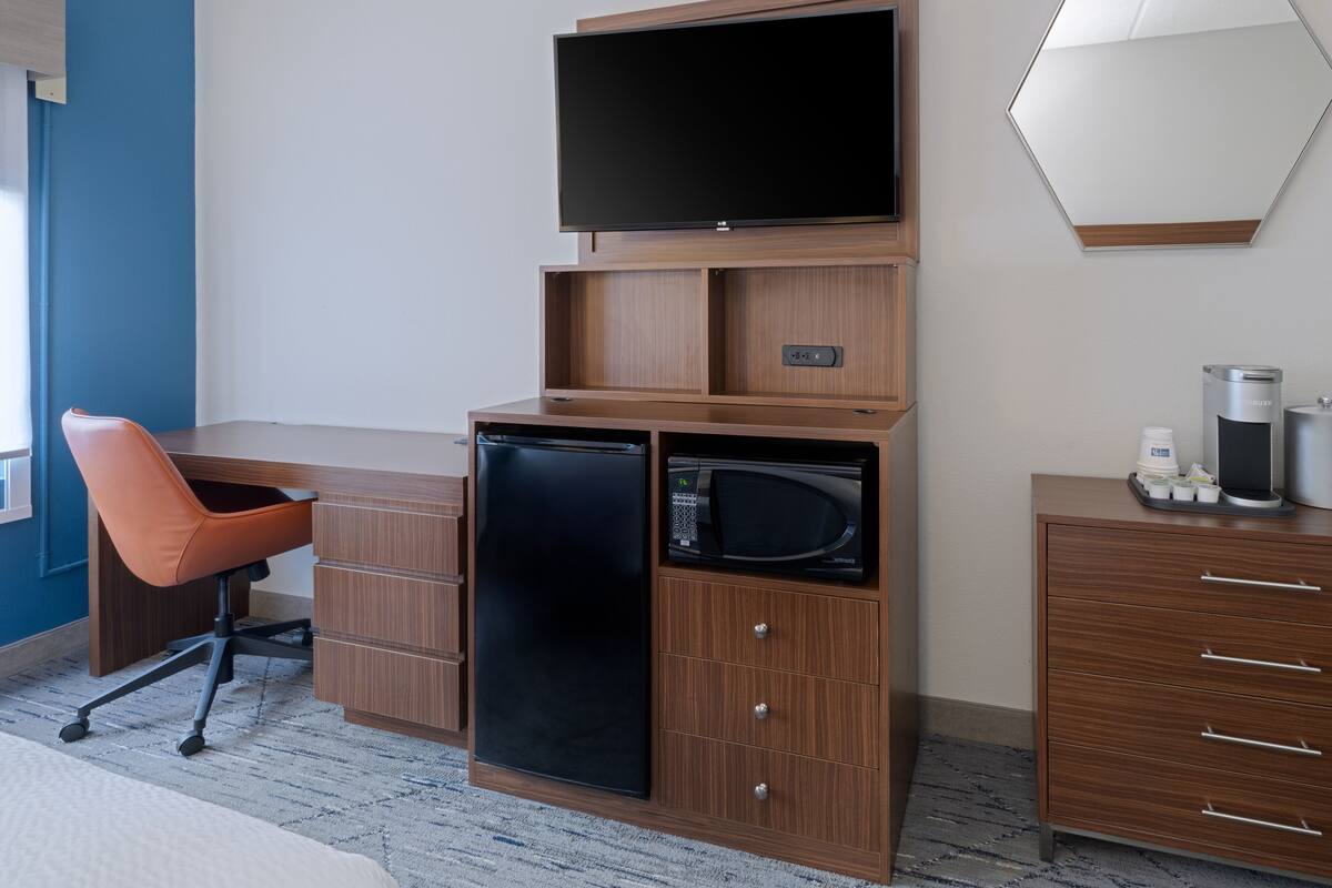 A hotel room with a wooden desk, a black refrigerator, a microwave, a television, and a coffee machine.