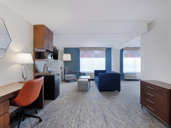 Une chambre d’hôtel moderne équipée d’un bureau, d’un fauteuil de bureau, d’un canapé et d’un pouf. Elle comprend deux fenêtres avec stores, un téléviseur à écran plat et une lampe.