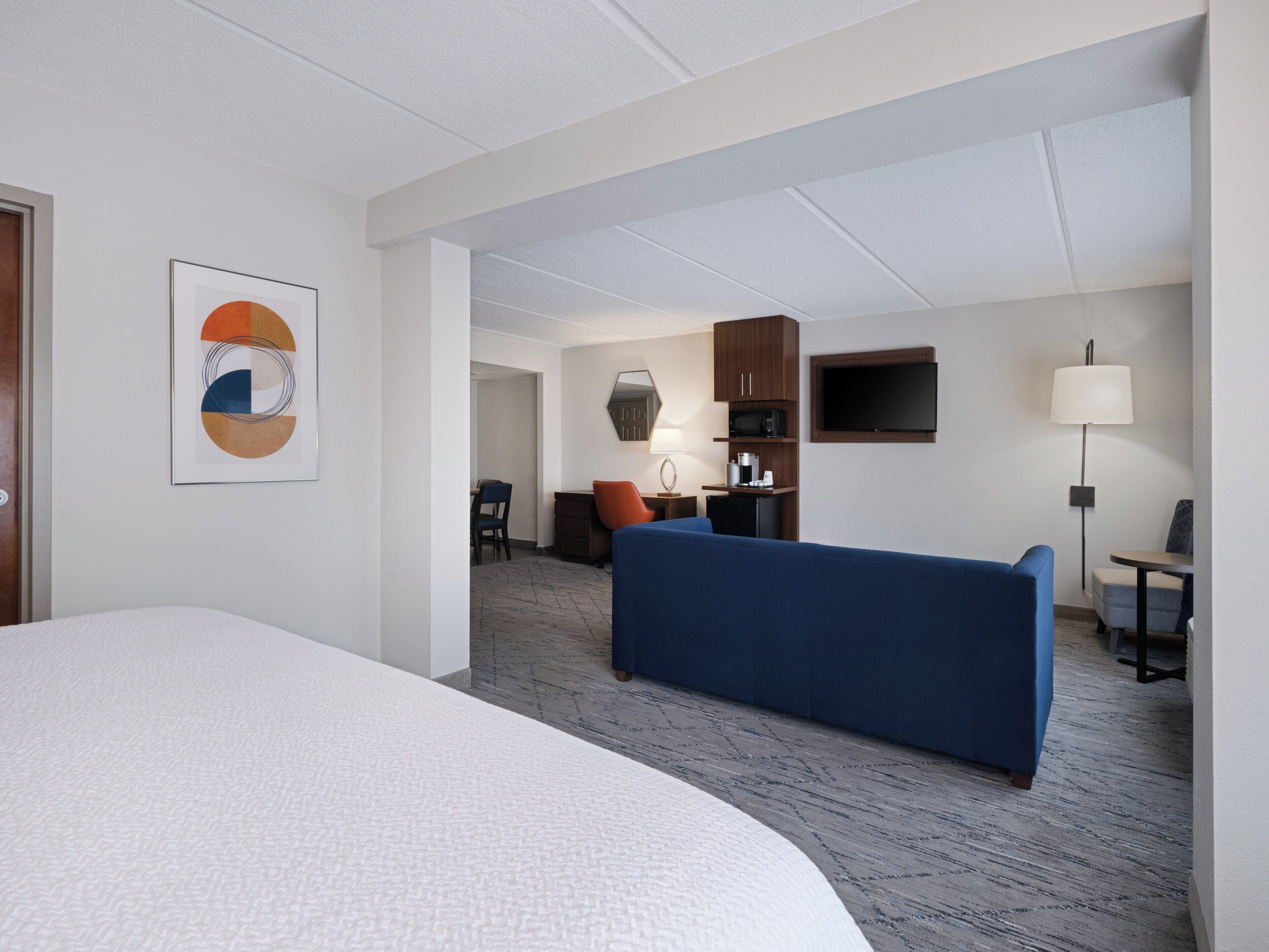 A hotel room with a large bed, a blue sofa, a desk with a lamp, a television, and a kitchenette.