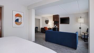 A hotel room with a large bed, a blue sofa, a desk with a lamp, a television, and a kitchenette.