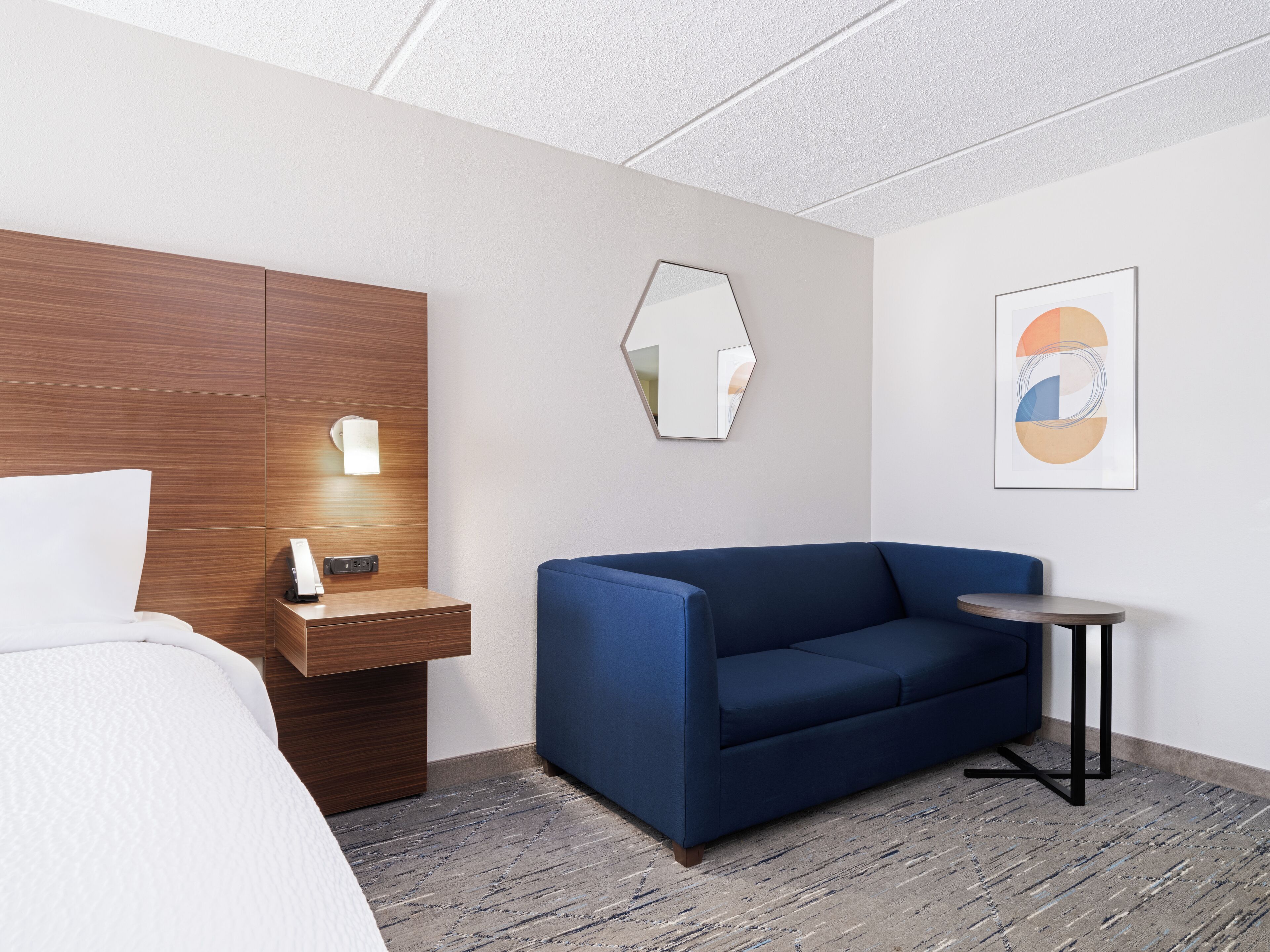A hotel room with a bed, a wooden headboard, a blue sofa, a small round table, and a wall-mounted mirror.
