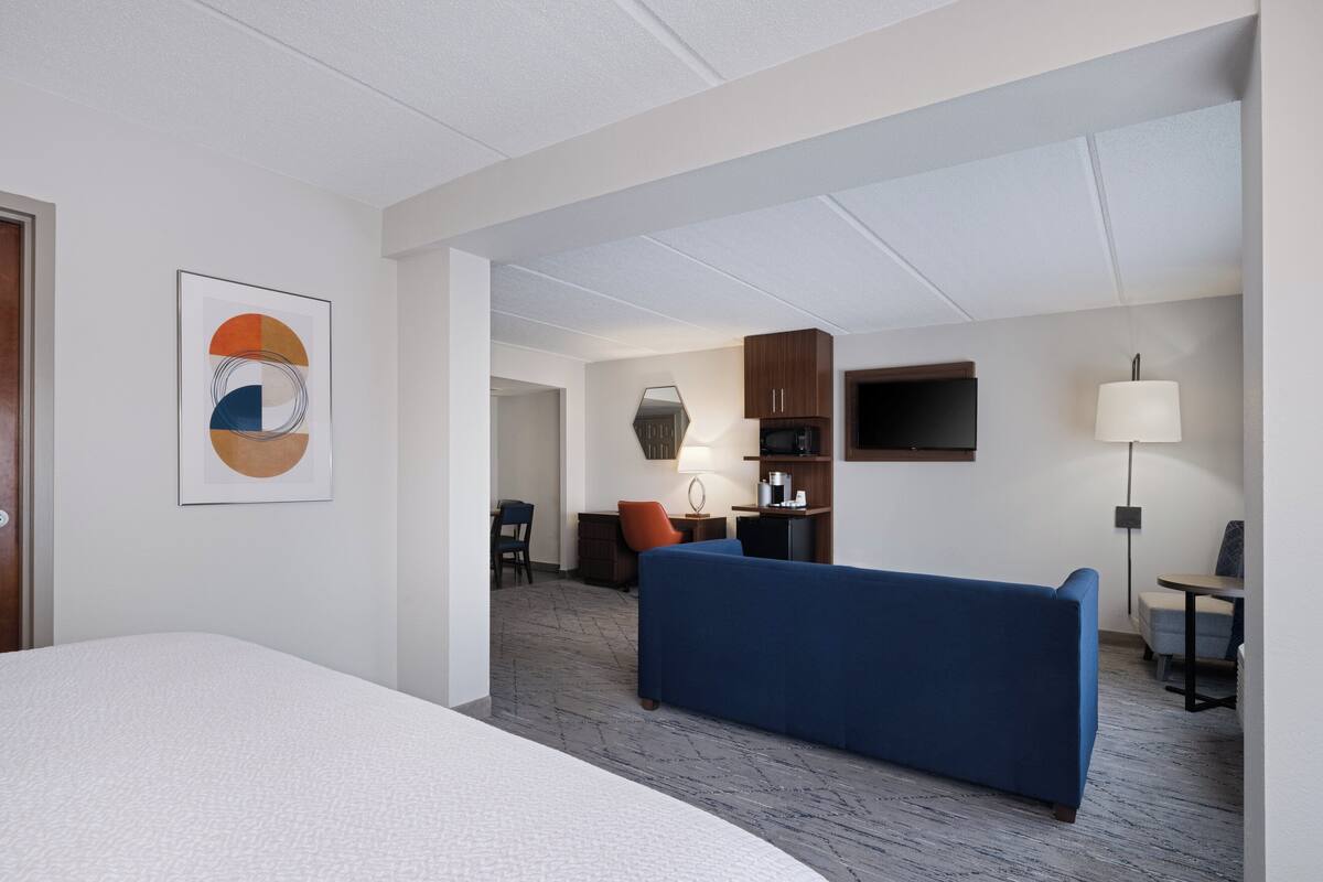 A hotel room with a large bed, a blue sofa, a desk with a lamp, a television, and a kitchenette.