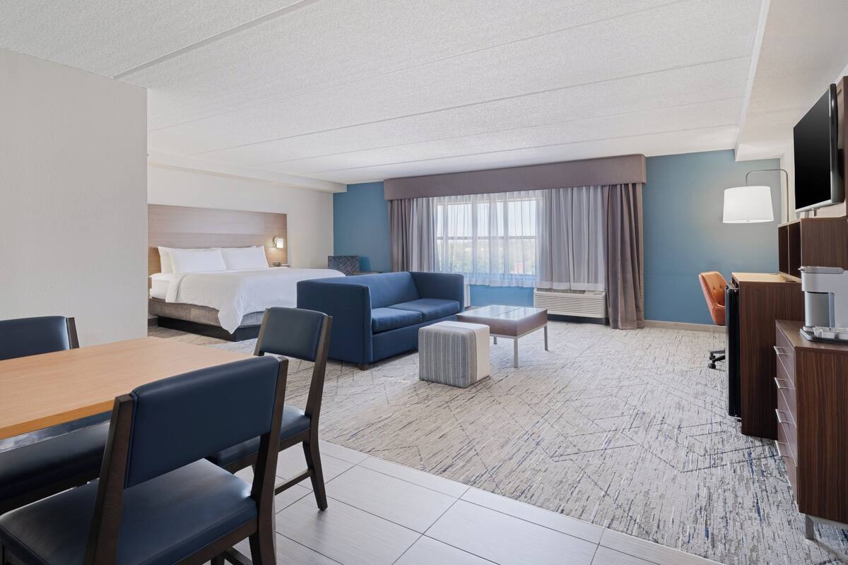 A hotel room with a bed, a sofa, a dining table, chairs, a TV, and a coffee machine.