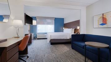 A hotel room with a bed, a desk, a chair, a sofa, a small table, a TV, and a window with blinds.