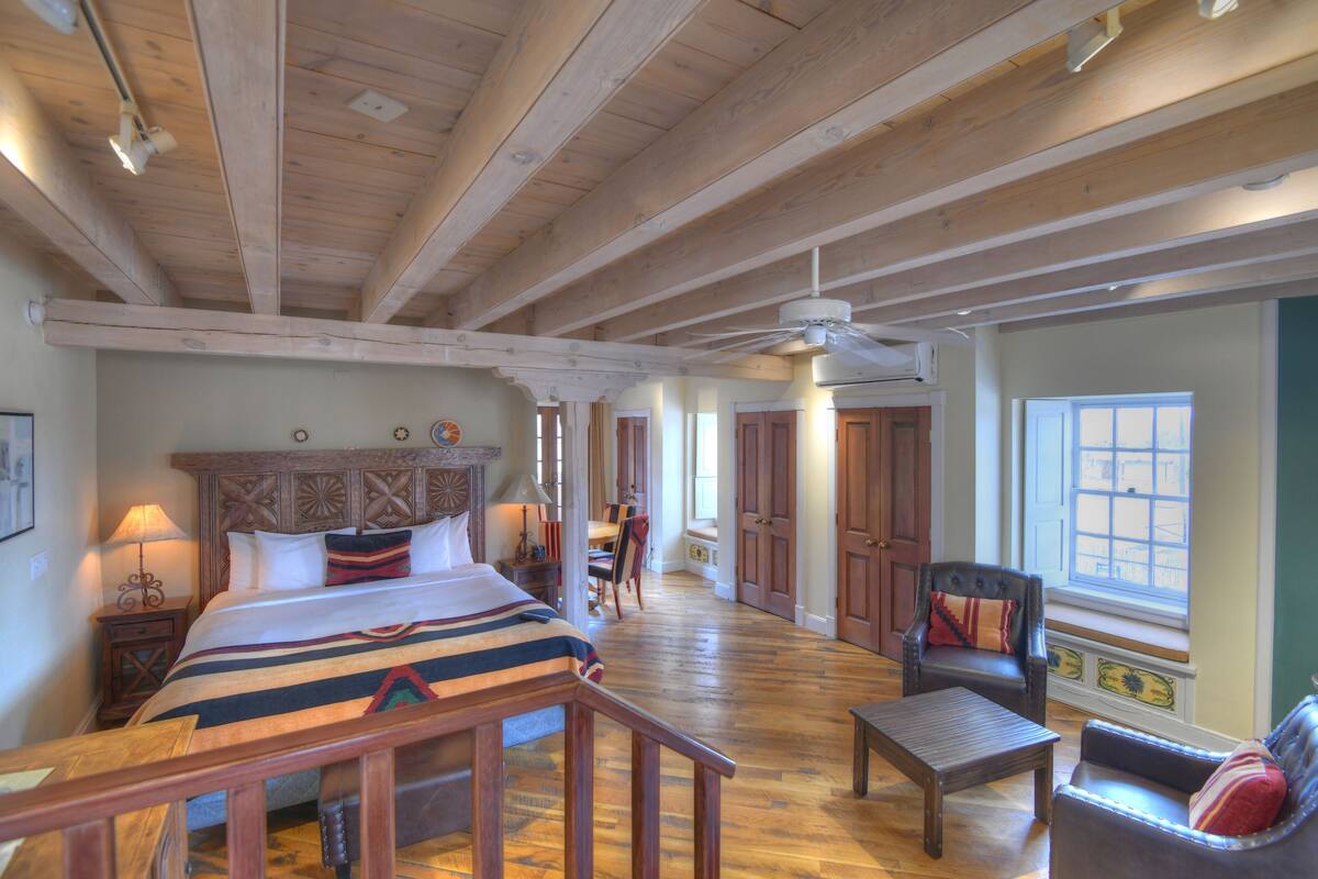 A spacious room with a large bed, wooden ceiling beams, a ceiling fan, and a view of a window.