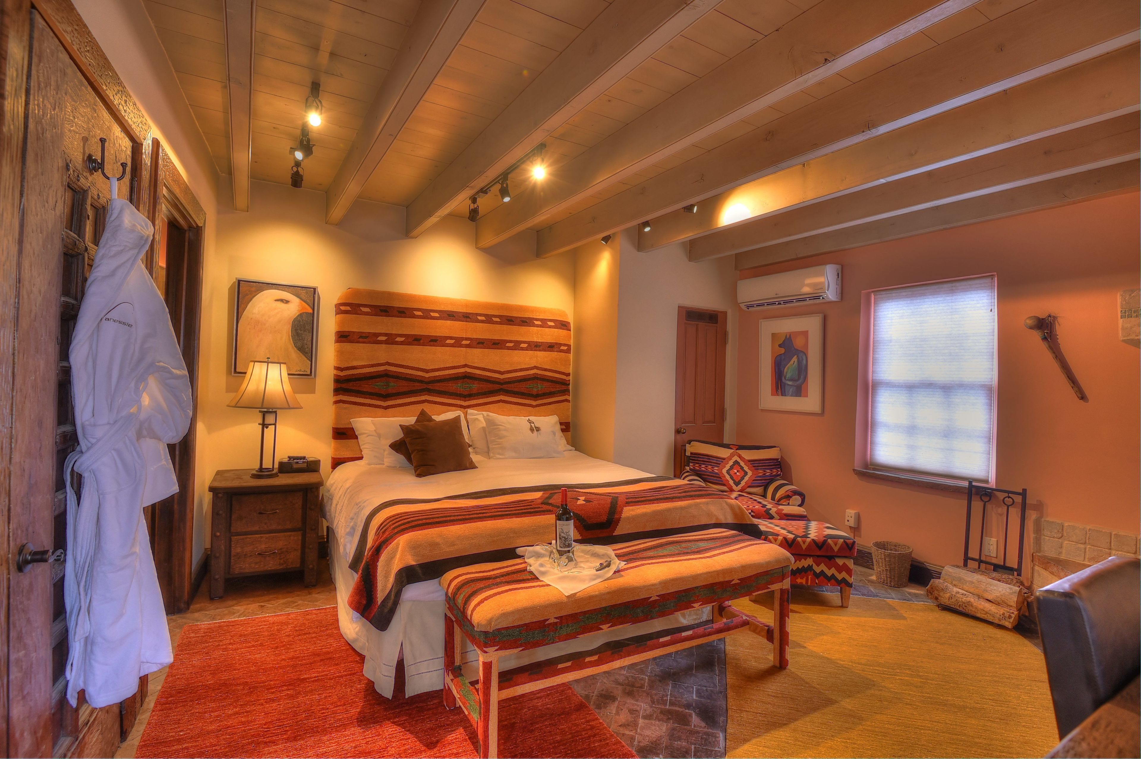 A cozy bedroom with a wooden ceiling, a large bed with patterned bedding, a bench, a robe hanging, and a window with blinds.