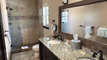 A bathroom with a granite countertop, two sinks, a large mirror, and a shower area.