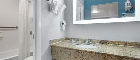 A bathroom with a granite countertop sink, a white toilet, a shower with a white curtain, and a security camera.