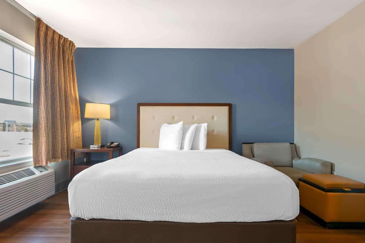 Habitación de hotel con una cama grande, una mesita de noche con lámpara, una silla y vistas a la ciudad por la ventana.