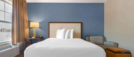 A hotel room with a large bed, a nightstand with a lamp, a chair, and a view of the city through the window.
