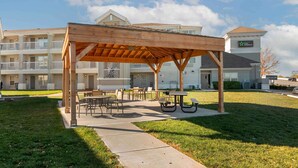 Un área de estar al aire libre con techado, mesas y sillas, una mesa de picnic y un edificio con el letrero Stay America al fondo.