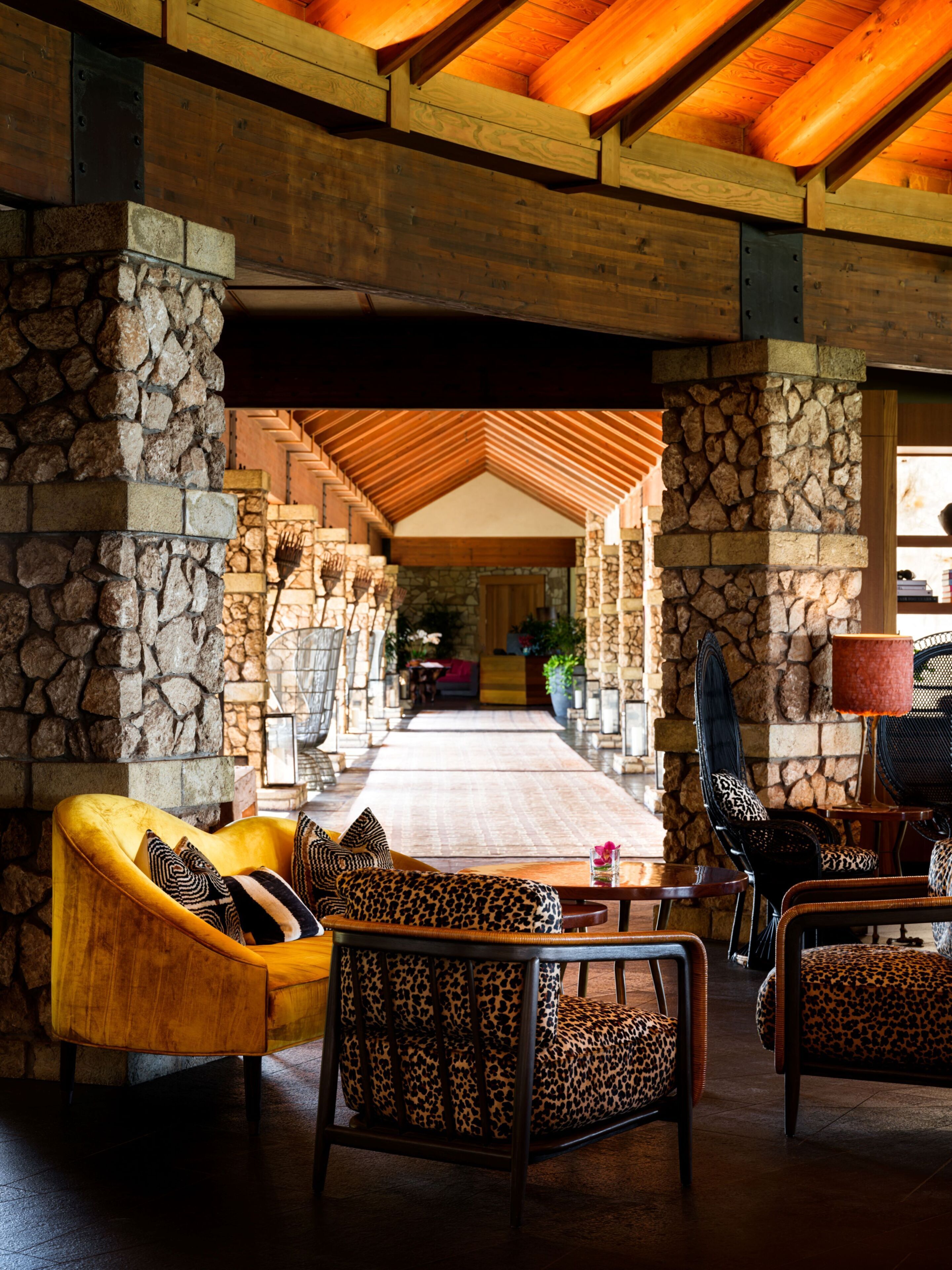 A spacious lobby with a stone wall, wooden ceiling beams, and a central seating area with a yellow sofa and leopard print chairs.
