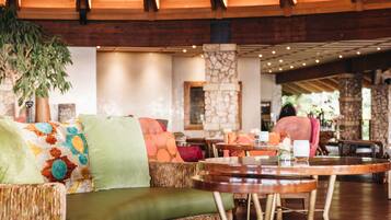 A spacious lobby with a high wooden ceiling, a large green sofa with colorful cushions, and a dining area with wooden tables and chairs.