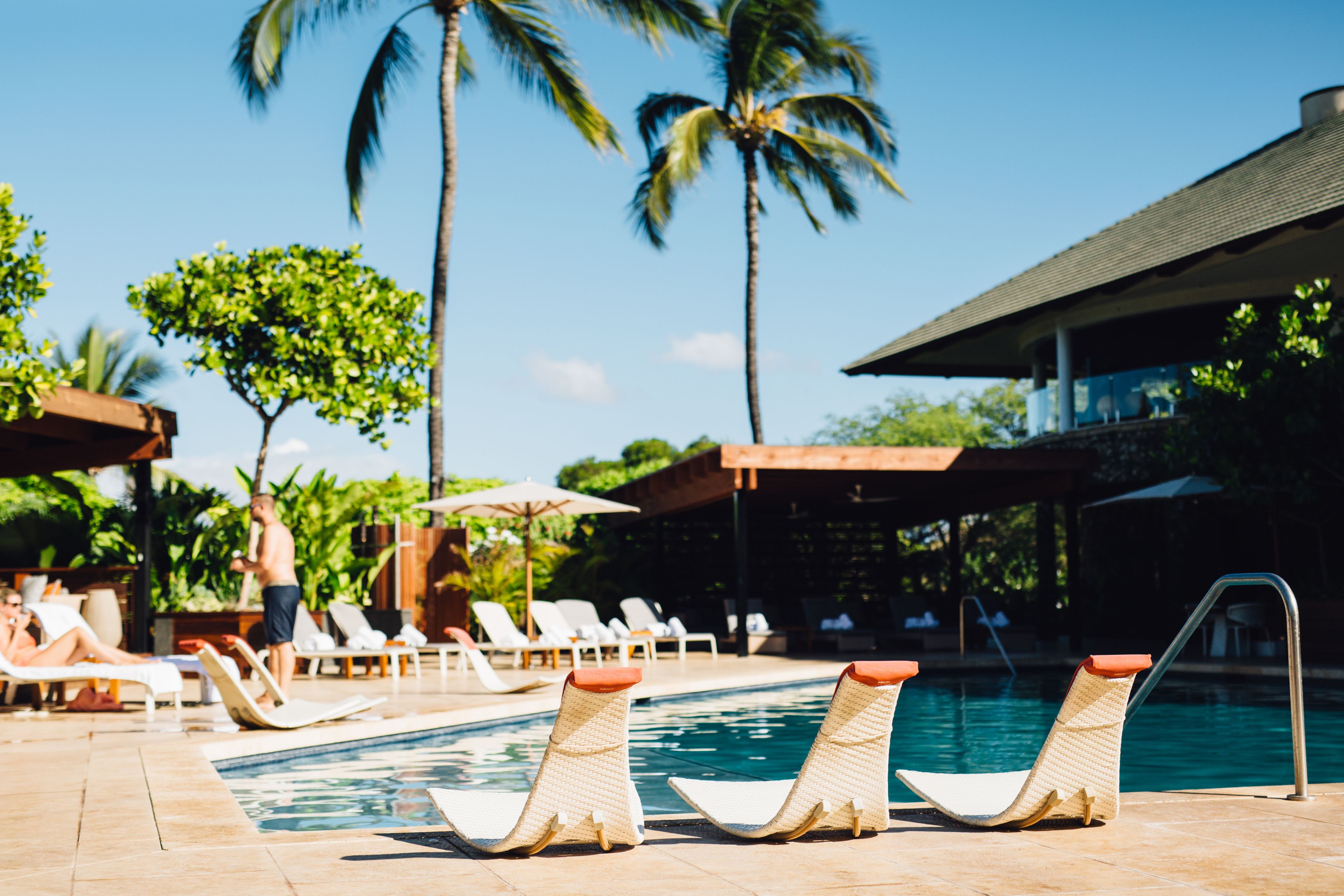 A poolside area with lounge chairs, a swimming pool, palm trees, and a covered seating area.
