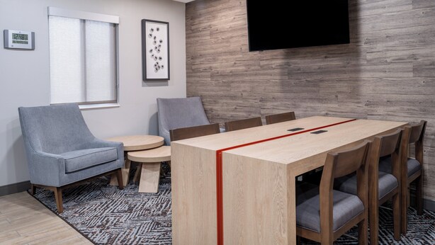 Una sala de reuniones moderna con una mesa de madera, sillas grises, un televisor montado y un reloj en la pared.