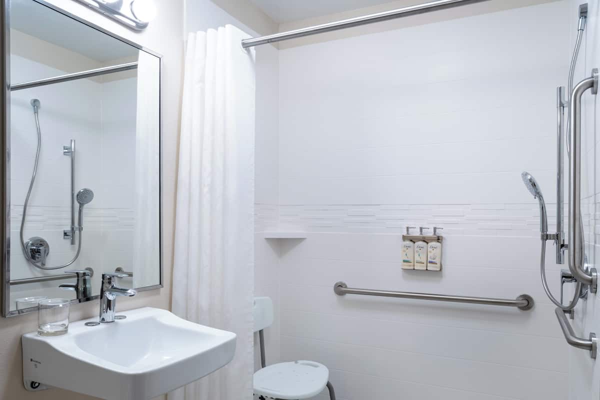A modern bathroom with a white sink, a handheld showerhead, a glass shower enclosure, a small white stool, and a handrail.