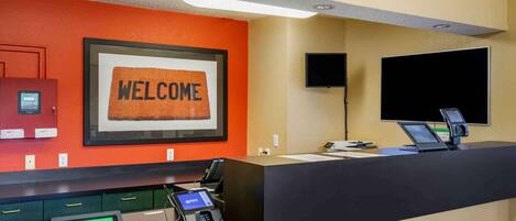 A hotel reception area with a 'WELCOME' sign, a reception desk, a flat-screen TV, and a digital kiosk.