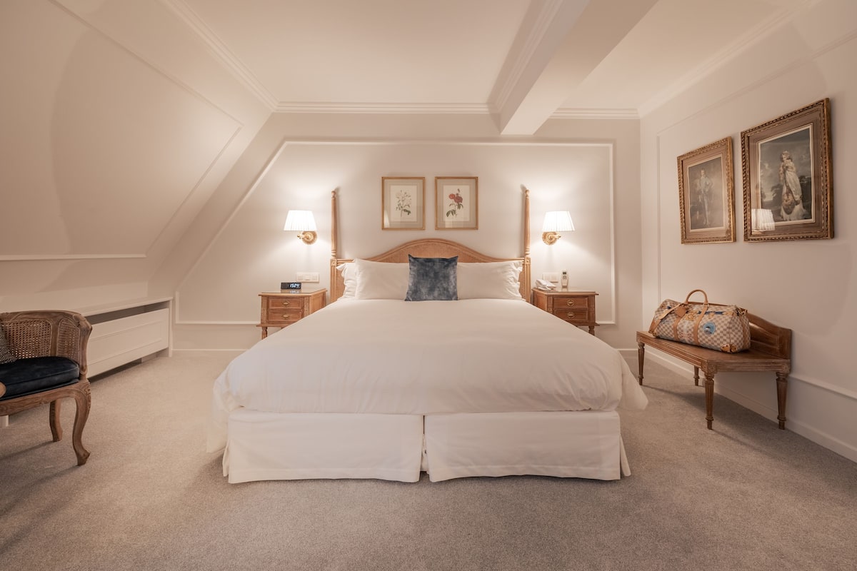 Une chambre spacieuse avec un grand lit, deux tables de chevet avec des lampes, un banc en bois et des tableaux encadrés aux murs.